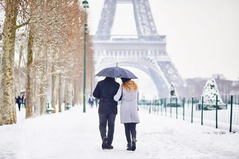 Couple walking along a tree-lined path toward the Eiffel Tower in the snow